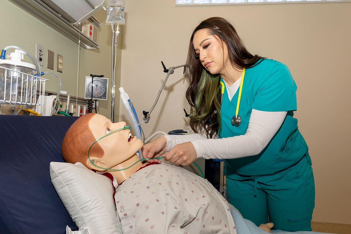 Student wearing scrubs adjusts equipment on training manikin.